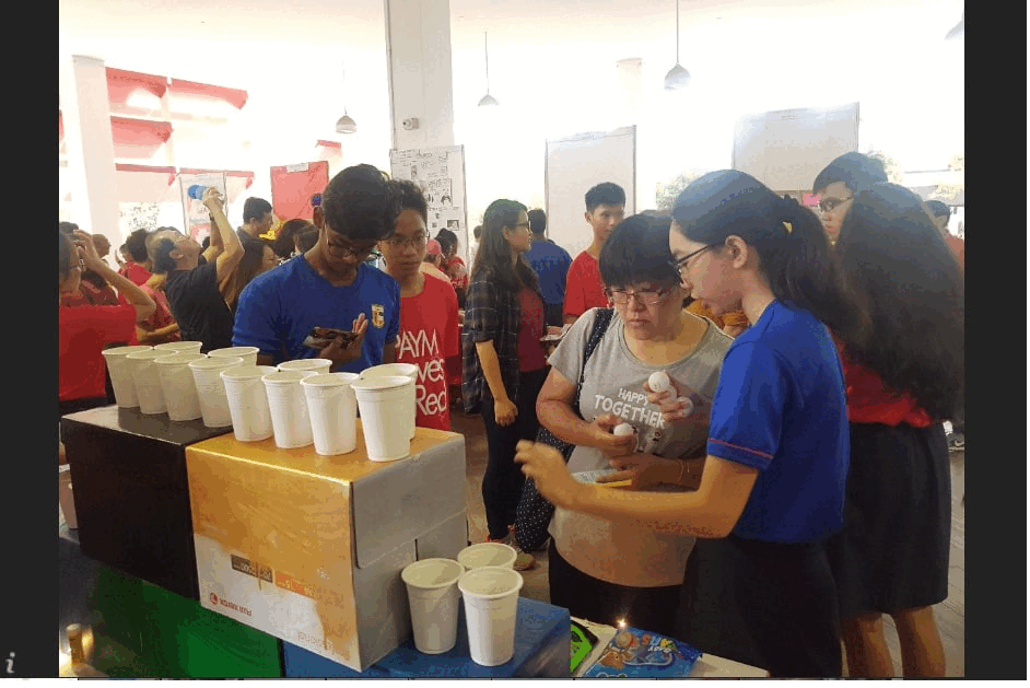 People in blue shirts stand near a table with rows of white cups at a busy indoor event.